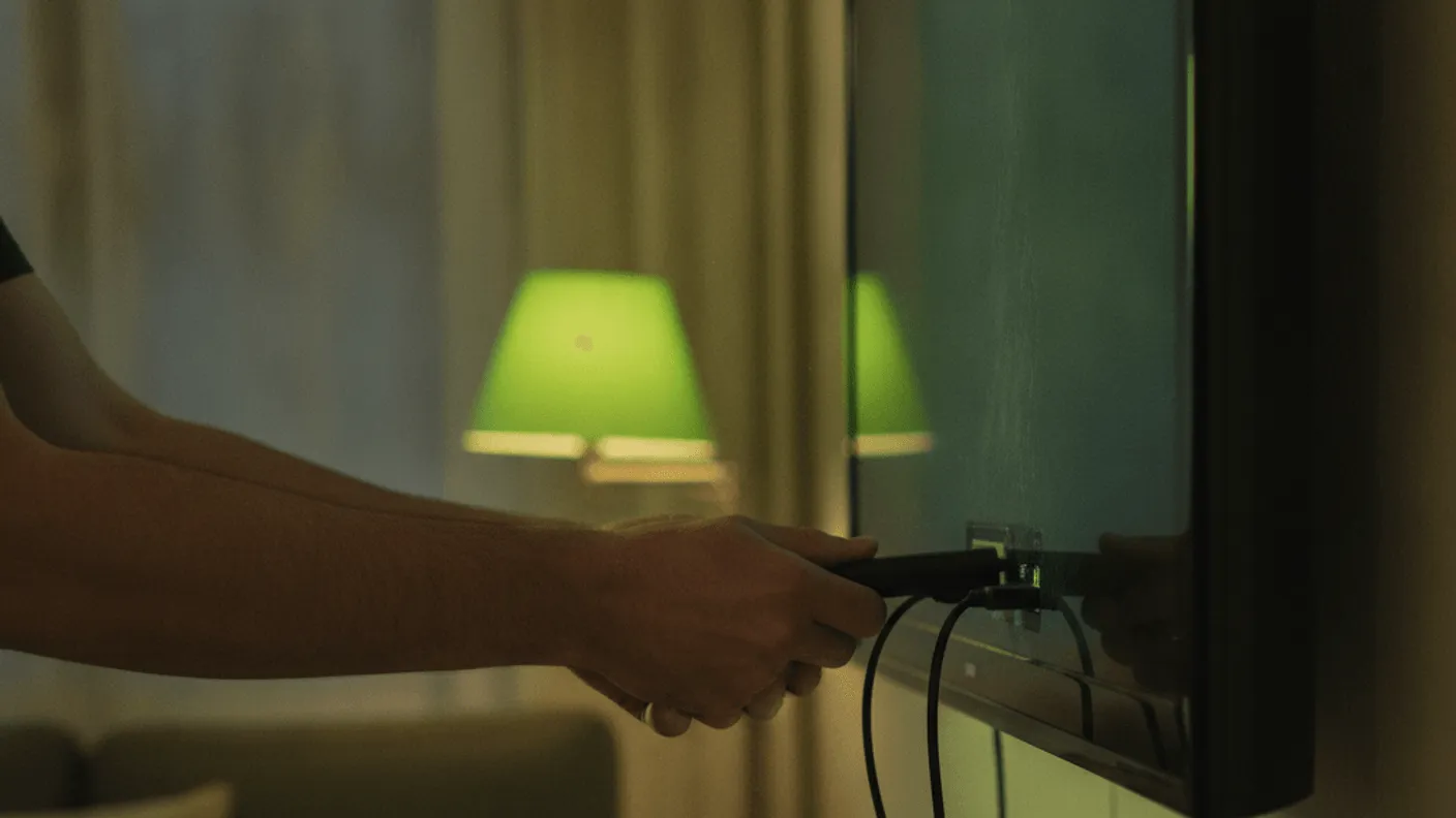 A man's hands plug an unbranded streaming device into the back of a wall-mounted TV in a dimly lit living room.