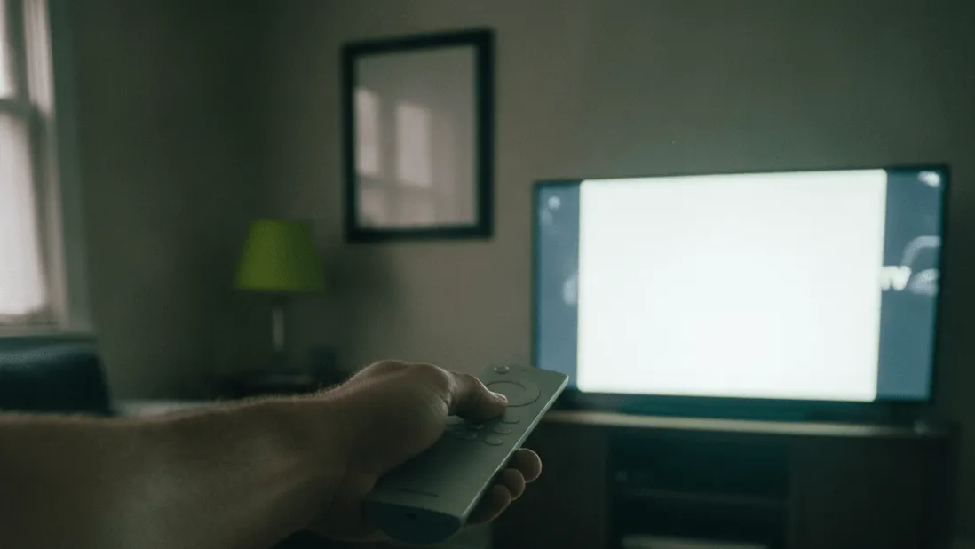 A hand holds a remote toward a television in a dim living room, with a notebook on a coffee table in the foreground.