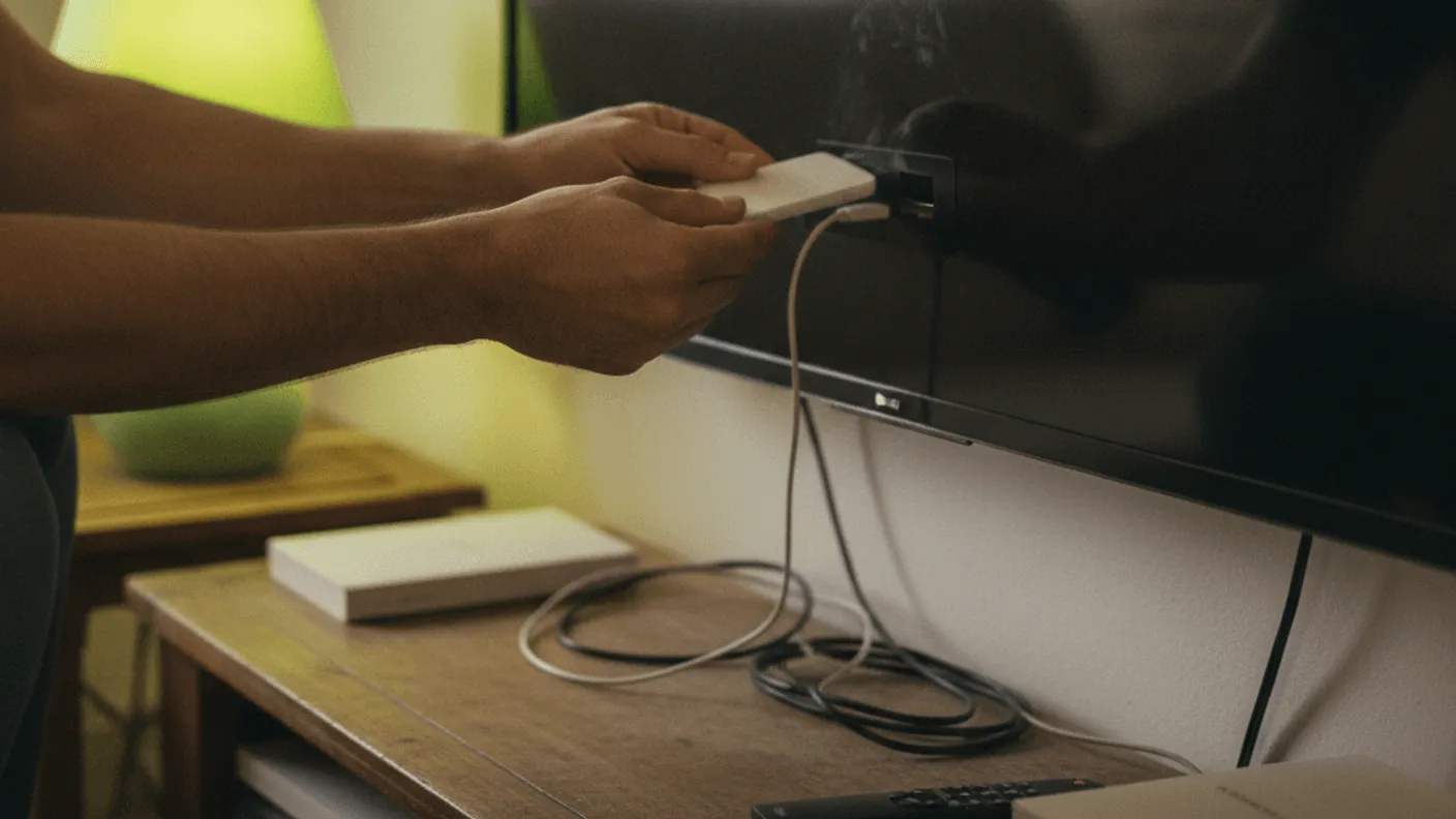 A person's hands plug a white streaming stick into the back of a wall-mounted television in a warmly lit room.