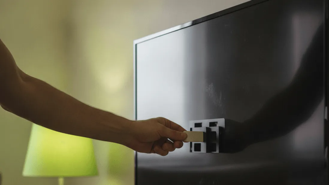 A hand plugs a small stick device into the back of a wall-mounted television in a warm, lived-in room.