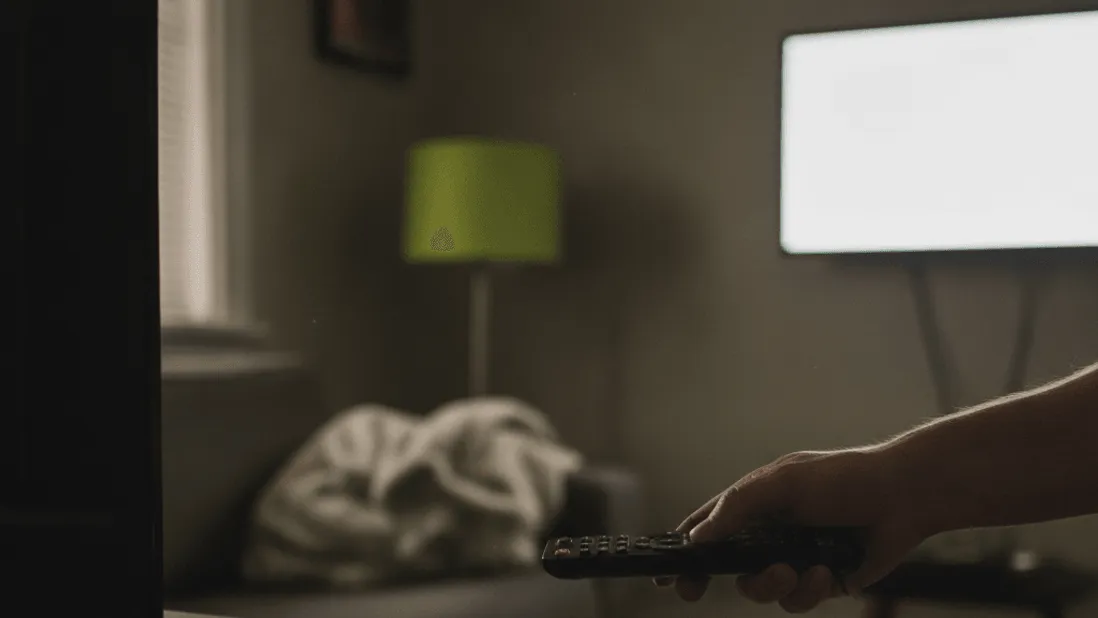 A hand holds a remote control near a media box with a green light in a dim room.