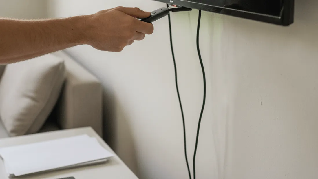 A hand inserts a streaming stick into the back of a wall-mounted television, with cables and a lime green glow.