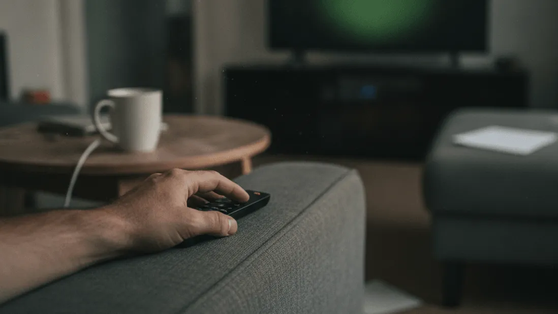 A man's hand rests a black remote on a grey sofa arm with a coffee mug and blurry TV in the background.