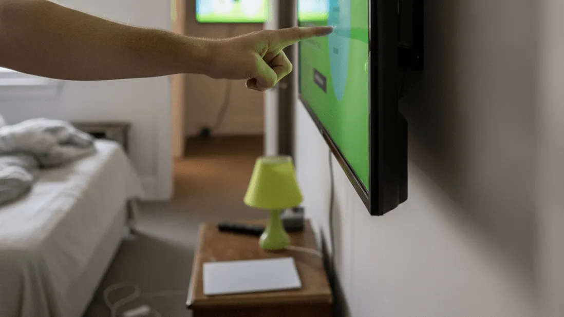 A hand adjusts a wall-mounted Smart TV in a bedroom, with another television visible through a doorway.