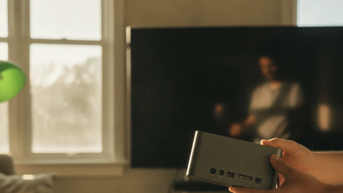A person's hands hold a streaming box, inspecting its ports, with cables and a notepad on a sunlit desk.