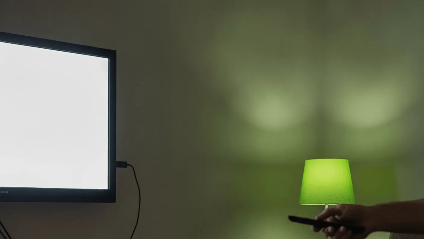 A person's hand holds a remote control in a dimly lit room with a television and a router.