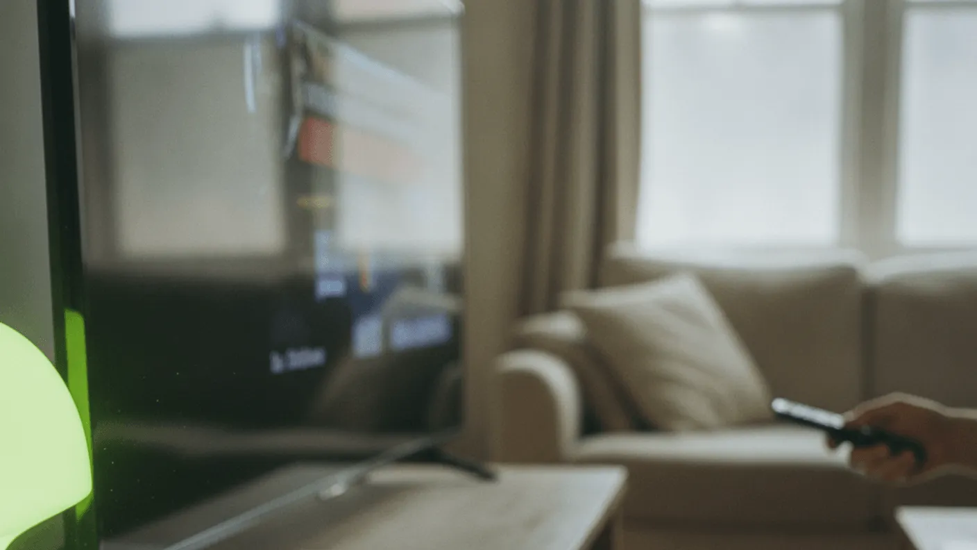 A small black streaming box sits on a wooden TV stand with a lime-green lamp, a TV, and a blurred hand holding a remote in the background.
