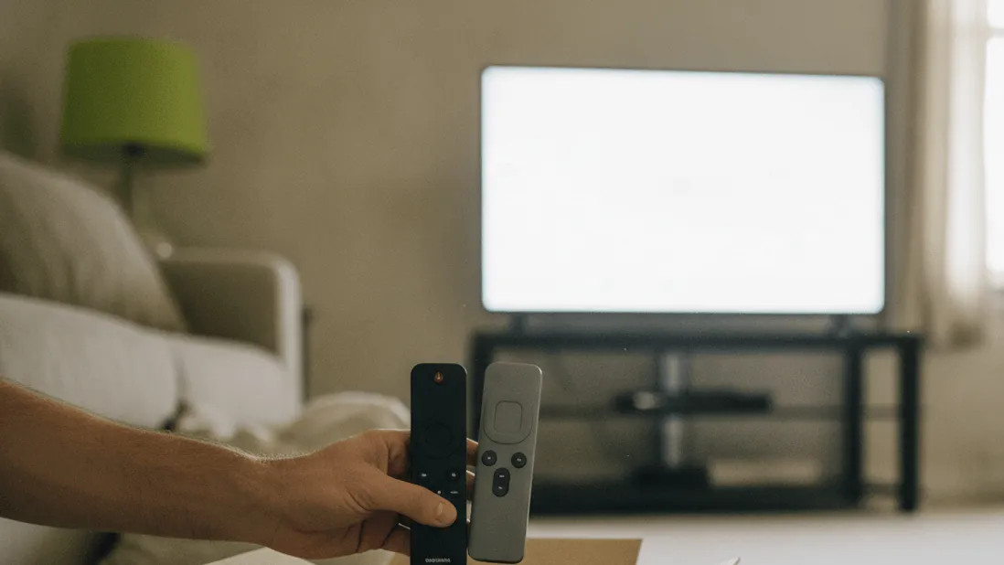 A hand holds two streaming remotes above an open cardboard box on a carpeted floor.