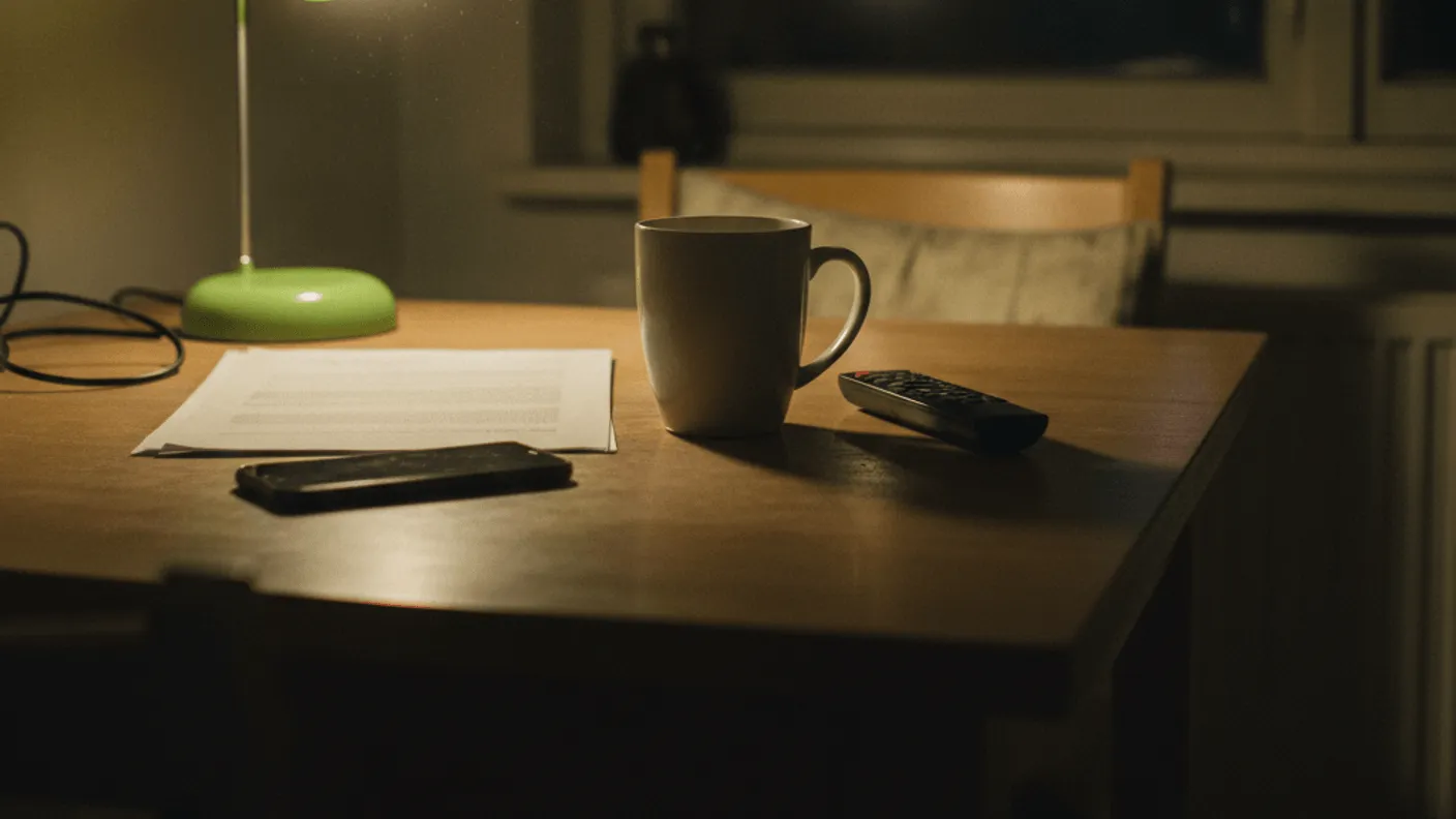 A kitchen table at night with a smartphone, paper, coffee mug, remote, and a lamp with a lime green cast.