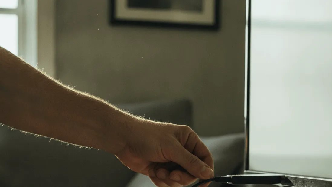 A man's hand inserts a streaming device into the back of a television, dust motes visible in daylight.
