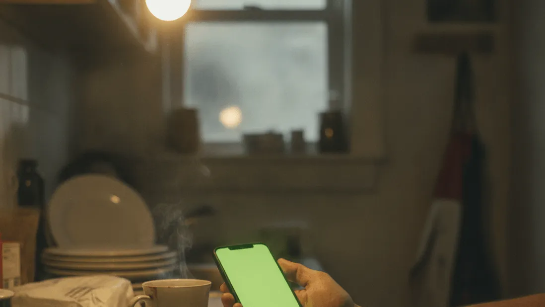 A hand holds a smartphone with a blank green screen next to a steaming mug on a kitchen counter in the evening.