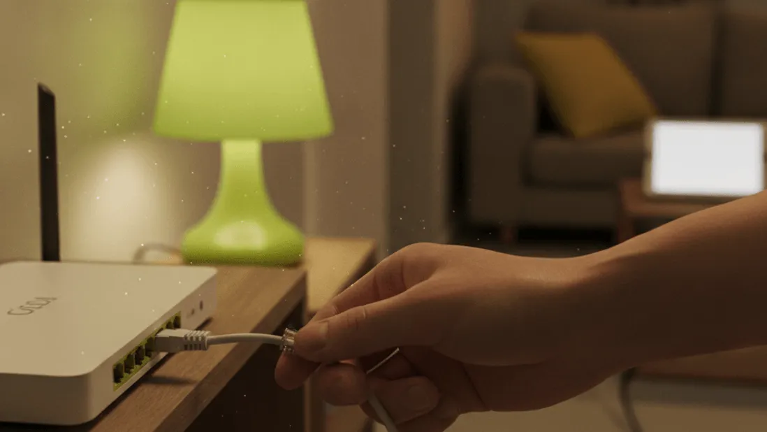 A hand holds an Ethernet cable near a router on a hallway shelf, evening light from a lime green lamp.